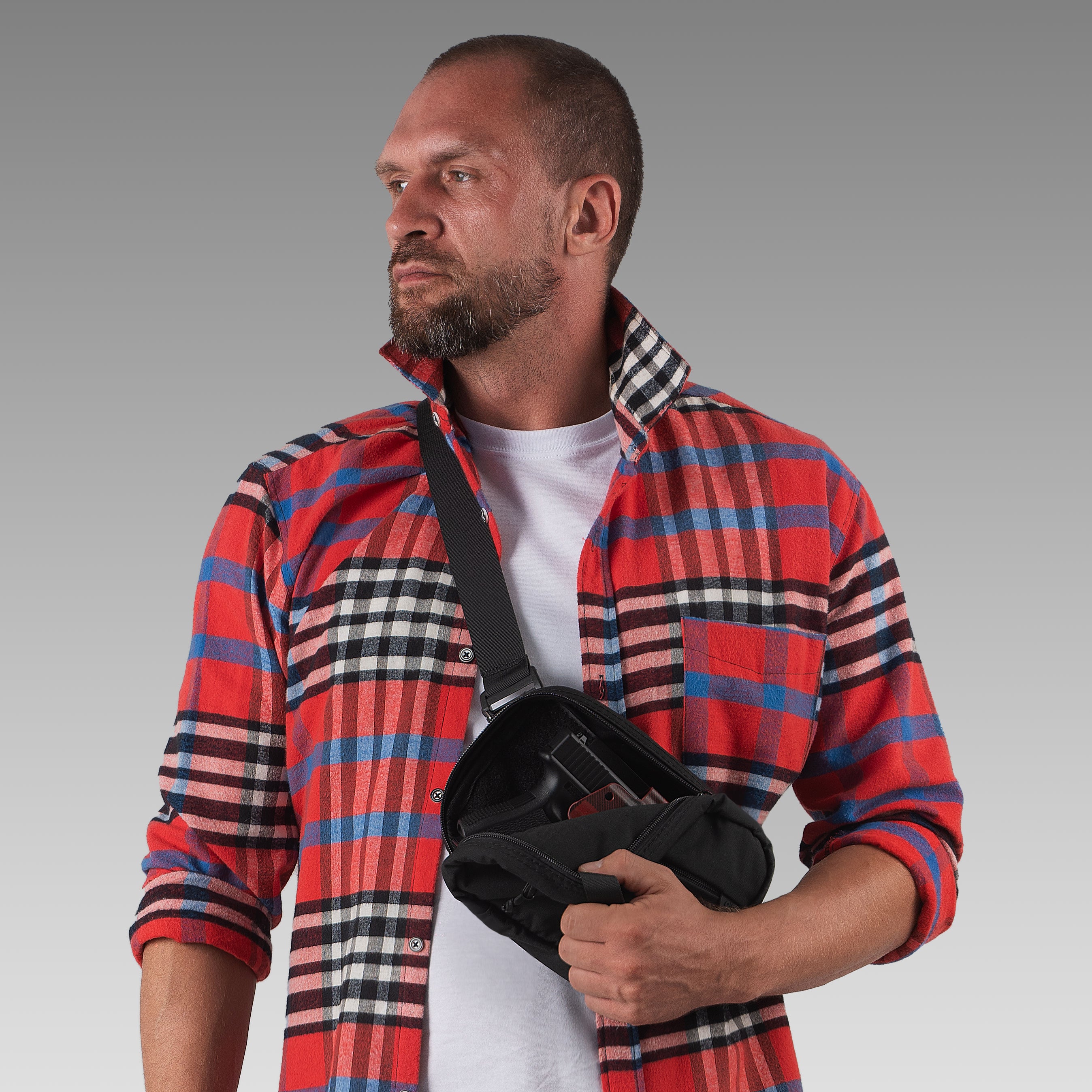 Man wearing a red plaid shirt holding a black bag against a gray background