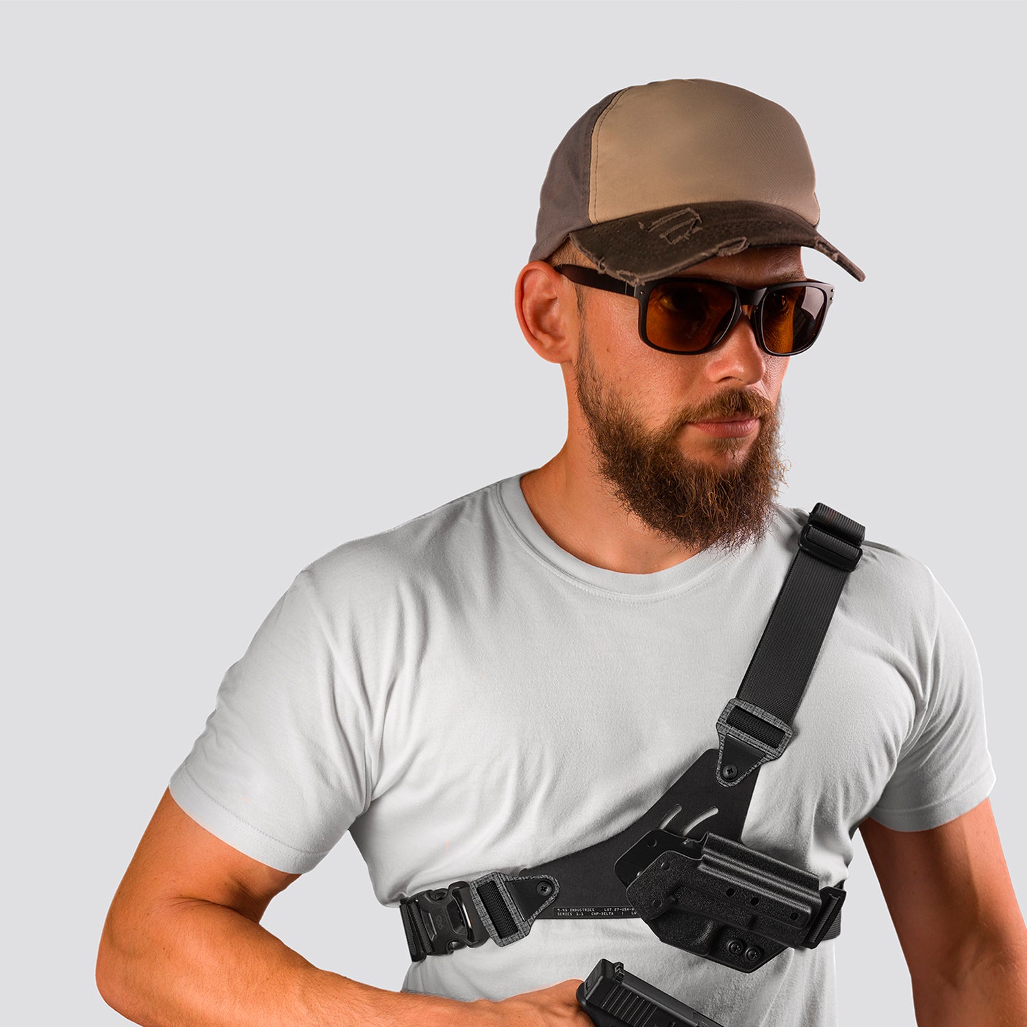 Man wearing a white t-shirt, brown cap, and sunglasses with a black shoulder holster on a light gray background