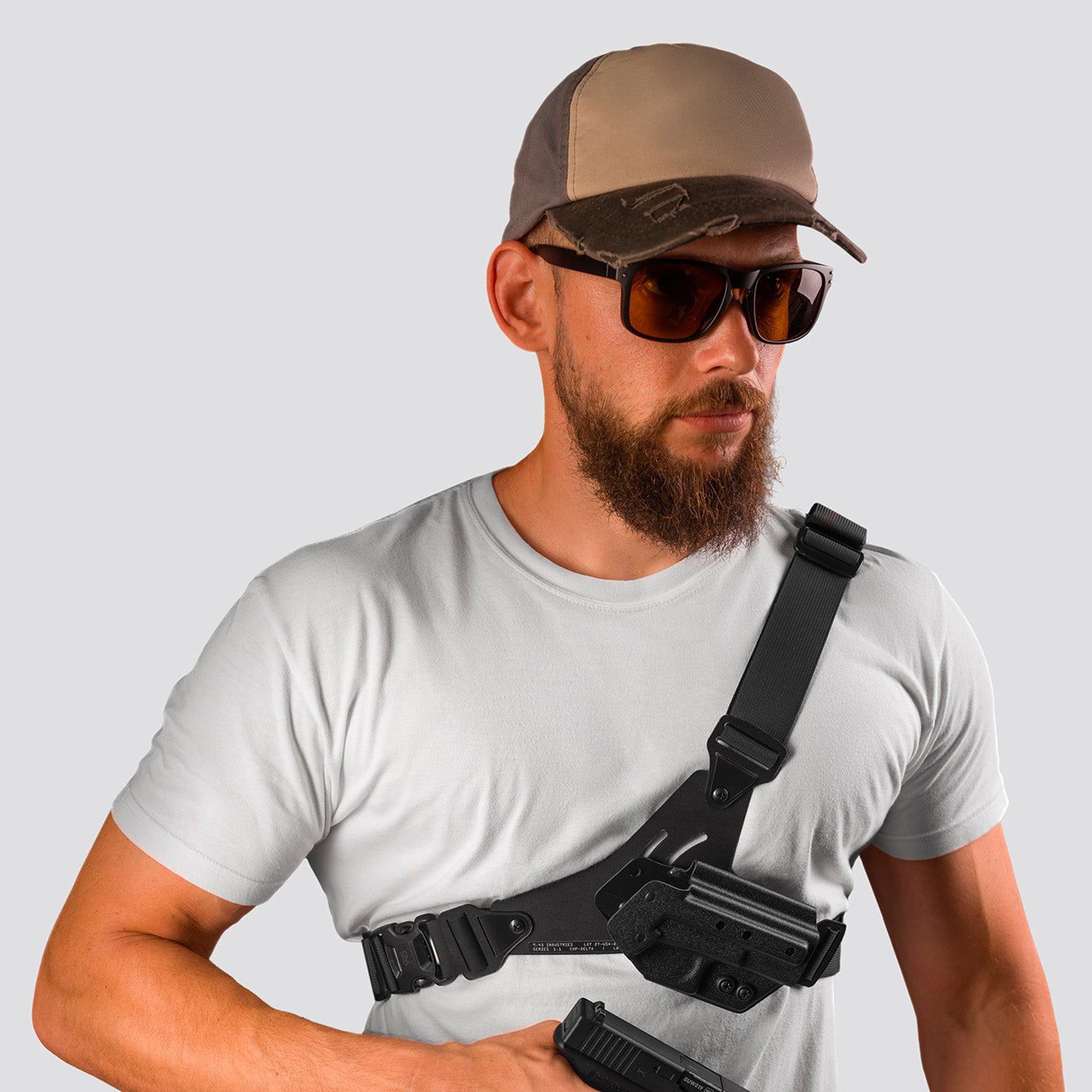 Man wearing a cap, sunglasses, and a white t-shirt with a black shoulder holster on a gray background